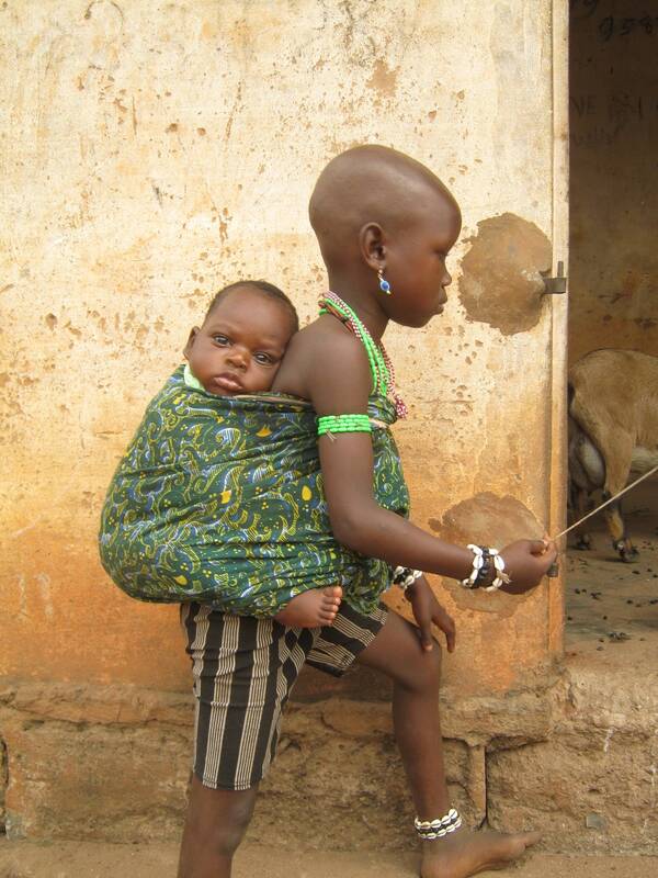 Young vodoun adept, shaved head and beaded bracelets, carrying a baby slung on her back, Benin